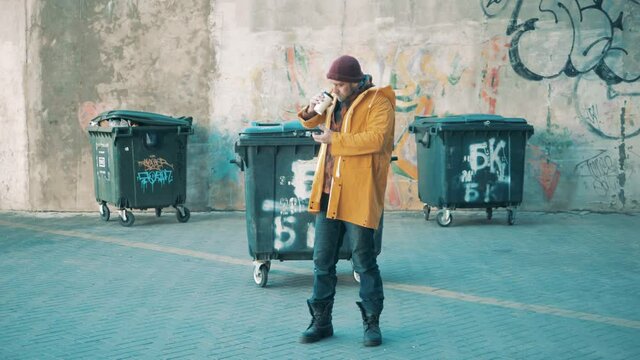 A poor man is drinking coffee next to the trash bins