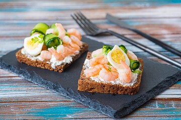 Smorrebrod, open sandwich with shrimps, creme cheese, boiled quail egg and cucumber slices on a slate or stone board on a wooden background. Sandwich recipes.