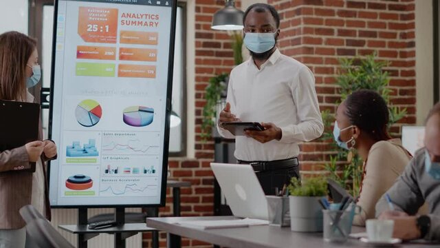 Man and woman with face mask planning business growth in front of coworkers at briefing meeting. Colleagues using monitor to do financial presentation for marketing strategy during pandemic. - Powered by Adobe