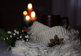 A cup of tea and a knitted scarf will give you warmth and comfort. The composition is decorated with a bouquet of small white flowers and a pine cone. Burning candles in the background. Close-up.