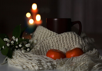 A cup of tea and a knitted scarf will give you warmth and comfort. The composition is decorated with a bouquet of small white flowers and two tangerines. Burning candles in the background. Close-up.