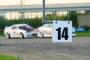 Close-up of the plate with the number 14. In its background, in out-of-focus two drift racing cars....