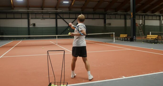 A man trains in a tennis court alone, throwing tennis balls over the net