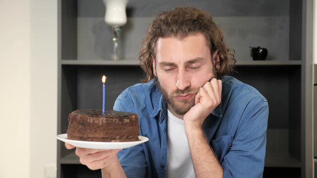 Nervous angry lonely man holds a cake with a candle and celebrates his birthday himself. Ruined celebration. Holiday loneliness. Festive killjoy. Sad annoyed disappointed loser man.