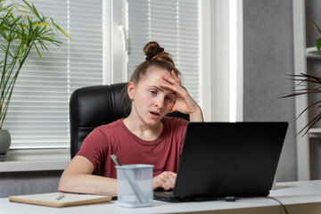 Young woman freelancer works from home. Girl sits at the workplace, works at laptop, tired of...