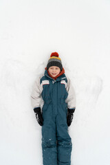 Fototapeta na wymiar Boy in a warm jumpsuit and hat lies in the snow. Winter holidays. Child is playing in the snow. Top view