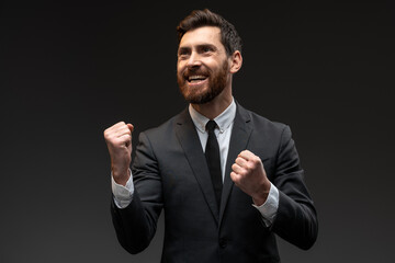 Portrait of happy screaming handsome bearded man in formal suit standing with excited face and rejoicing his victory. Indoor studio shot isolated on black background