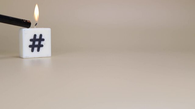 A square candle printed with a hash tag being lit and blown out on a brown background