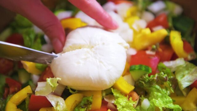 Macro closeup of hand knife person cutting burrata mozzarella Italian cheese ball with creamy center inside on fresh caprese salad with yellow bell peppers, tomatoes crumbled 