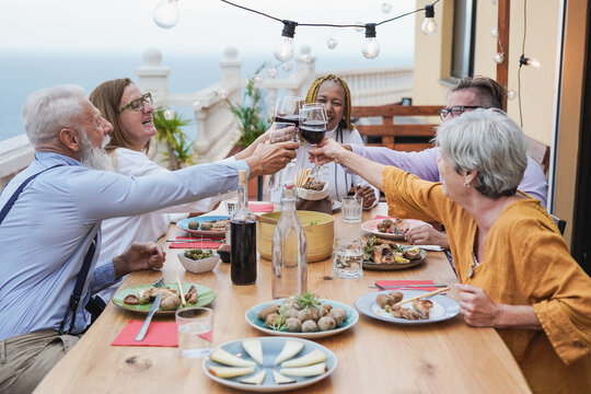Elderly friends cheering at dinner on patio at home - Multiracial senior people eating together