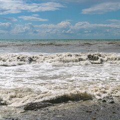 stormy waves of the Mediterranean sea run over the rocky summer beach against the backdrop of a blue cloudy sky