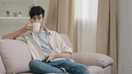 Young arabic man with glasses guy millennial arab hispanic sits at home couch with cup of tea coffee switches channels with remote control choosing movie program watching TV relaxing indoors weekend