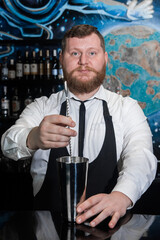 The hands of a professional bearded, adult bartender interfere with a bar spoon the contents of a metal shaker tool for preparing and stirring alcoholic cocktails