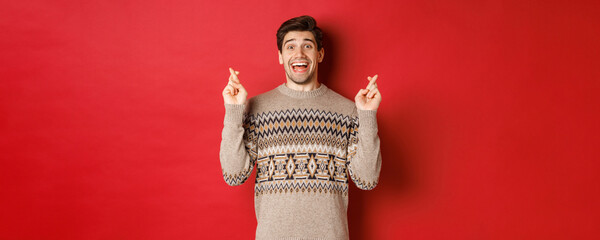 Image of excited and hopeful young man making wish, wearing winter sweater, crossing fingers for...