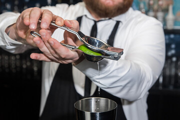Hands of a professional bartender squeeze lime juice with an iron tool with a juicer into a metal shaker, close-up
