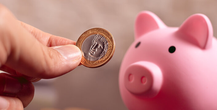 Real - BRL, dinheiro do Brasil. Um homem segurando uma moeda de 1 real com um cofrinho na composi&ccedil;&atilde;o em ambiente escuro. Conceitos de economia brasileira, finan&ccedil;as e poupan&ccedil;a.
