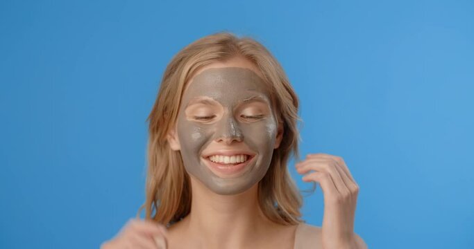 Blonde woman poses in applied clay face mask and smiles. Women's cosmetics usage. Beauty care on the blue screen, 4k Prores HQ