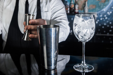 The hands of a professional bartender pour syrup into a measuring glass of jigger in a metal tool for preparing and stirring alcoholic cocktails of shaker drinks, and glasses with ice cubes