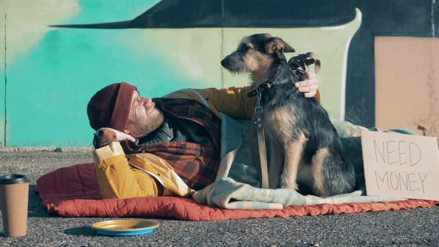 A poor man is stroking his dog while lying on the ground