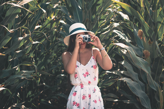 Outdoor lifestyle portrait of a young brunette girl taking photos with a vintage photo camera wearing summer white flowery dress and sun hat