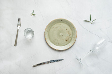 Green porcelain plate with stainless steel cutlery, glass vase with water and olive leaves on white marble table - Powered by Adobe
