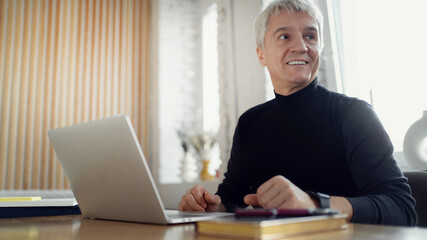 The man is a stylish freelancer, gray-haired workplace in the office. Works smiling on the internet on an online site.