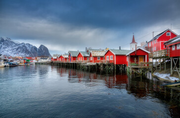 Beautiful red rorbu in overcast day. Fishing village in Lofoten islands, Norway. Winter landscape with houses, snowy mountains, sea, sky with clouds. Norwegian traditional red rorbuer on the water - obrazy, fototapety, plakaty