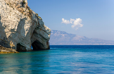 Cave in the rocky island cape in turquoise sea water with mountains on horison and a cloud on the blue sky