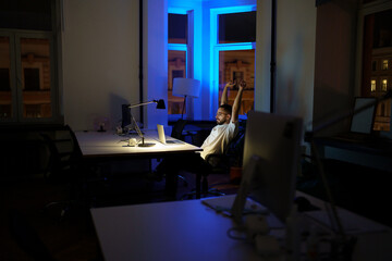 Male freelancer work late night stretching hands and back while sitting at laptop computer in dark office. Tired man programmer web designer overtime within deadline. Exhausted developer having break