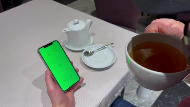 Male hand holds iPhone 12 Pro Max with chromakey green screen and drinks tea