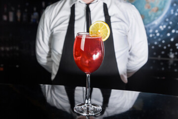 Red alcoholic beverage cocktail in glass decorated with a slice of lemon on the bar counter against the background of the bartender