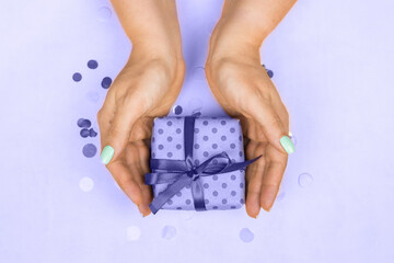 Woman's hands with blue pastel manicure holding a gift with a bow on a purple background very peri 2022. The concept of the holiday, gifts, surprises, birthday, new year and Christmas