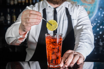 A professional bartender puts a slice of lemon in a red chilled alcoholic cocktail with bar tweezers in glass at a nightclub counter