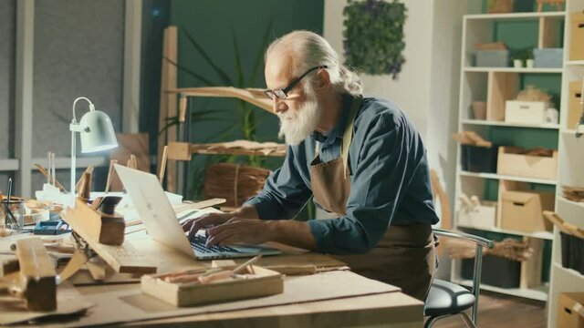 A Focused Carpenter Develops Layouts from Wood, Models 3D Sketches Using a Laptop in a Home Workshop. Garage Hobby, Profession, Joinery and New Computer Technology.