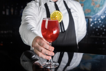 Professional bartender hand serves red alcoholic beverage in glass cocktail decorated with a slice of lemon on the bar counter