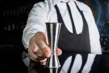 The hand of a professional bartender holds a jigger tool or measuring cup to control the ingredients added to the cocktail