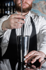 The hands of a professional bearded bartender interfere with a bar spoon the contents of a metal shaker tool for preparing and stirring alcoholic cocktails