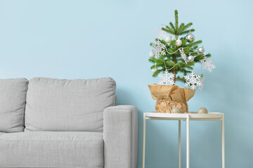 Beautiful Christmas tree in pot decorated with snowflakes, balls on table and comfortable sofa near light wall