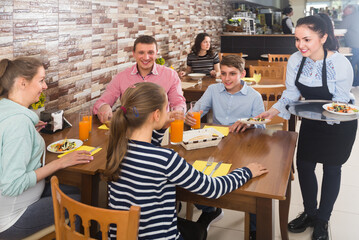 Polite smiling waitress bringing ordered dishes to family in cafe - Powered by Adobe