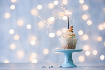 Stand with delicious Christmas cupcake and sparkler on table against blurred lights
