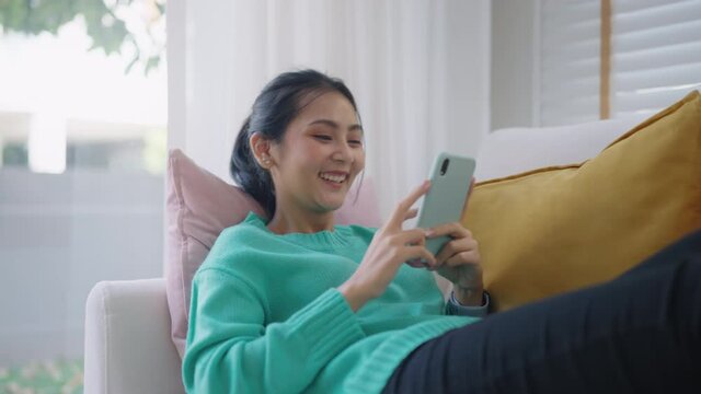 Zoom out asia young people generation z student casual teen girl enjoy fun look humor funny movie on TV app sit lying read text on cozy sofa play video live tiktok reel story with smile joy face.