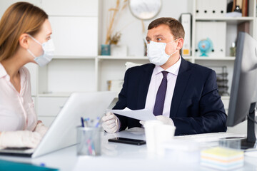 Fototapeta premium Portrait of busy entrepreneur in medical face mask and latex gloves working with female coworker in office. Concept of precautions and social distancing in coronavirus pandemic..
