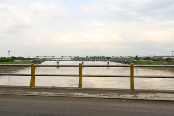 View of iron railway bridge above river from moving car in East Java, Indonesia with clouds in blue sky background. No people.