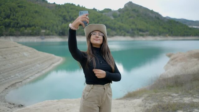 Girl with hat and glasses takes selfie and uses phone by lake in Spain