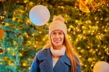 A smiling girl stands against the background of a large glowing Christmas tree. Selective focus. Festive mood