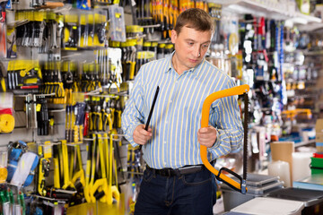 Confident glad man is choosing hacksaw in tools store - obrazy, fototapety, plakaty