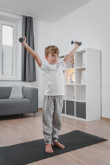 Fototapeta na wymiar Healthy lifestyle. Portrait of a boy exercising with dumbbells at home in the living room. The child is dressed in a white T-shirt and gray pants