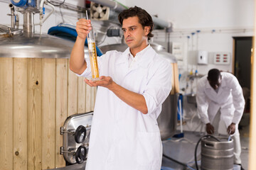 Young man expert is standing with flask for beer and analizying it in beer factory