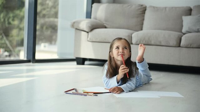Lies on the floor and draws. Cute little girl in nightwear is home alone at daytime in modern room