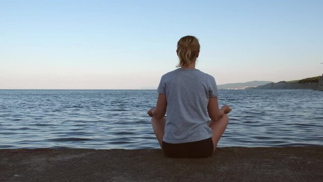Young woman sits in a lotus position on the pier and looks at the sea dawn of the sun. Sports, meditation, calmness, yoga, harmony, summer. Concept of nature, health, freedom, solo activity